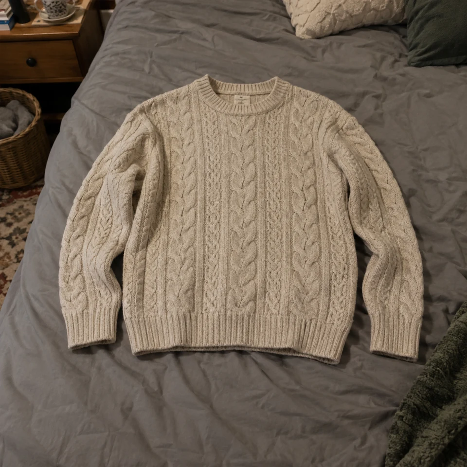 oatmeal cable-knit crewneck sweater with ribbed cuffs and hem, laid flat on a bed