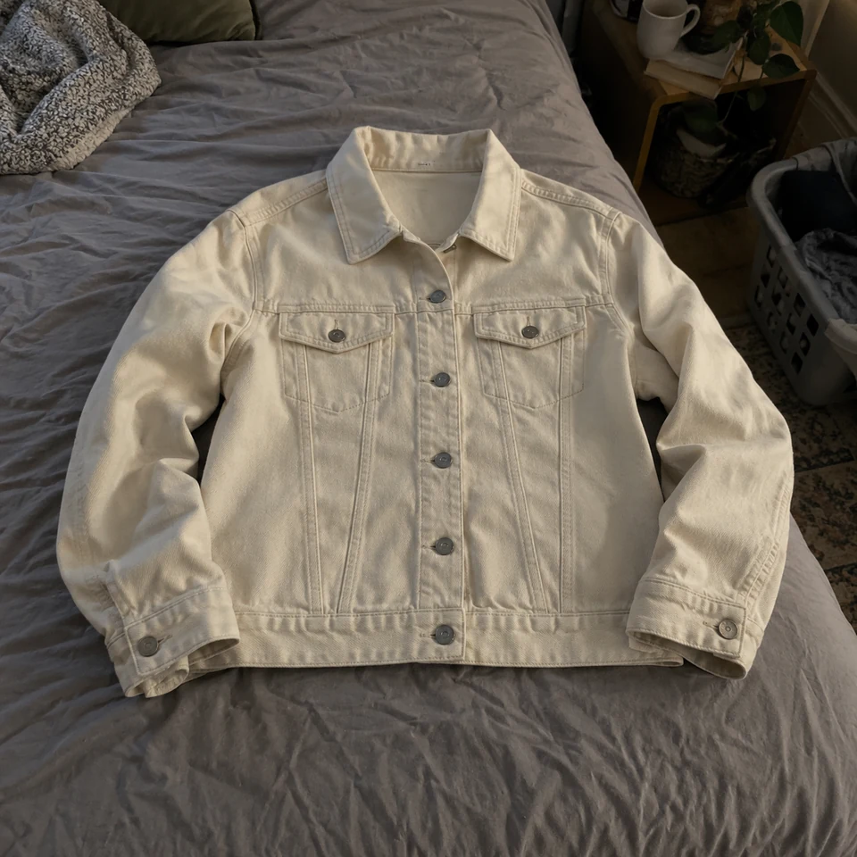 warm ivory denim jacket with six buttons and two chest pockets, laid flat on a bed