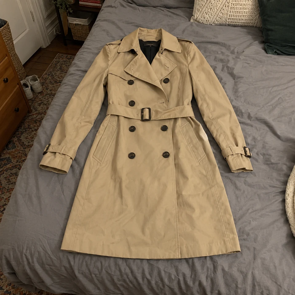 beige double-breasted trench coat with belt and dark buttons, laid flat on a bed